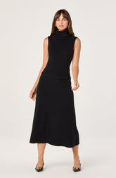 A woman is wearing a sleeveless, black turtleneck top paired with a matching long black skirt that reaches her ankles. She has on pointed-toe black shoes with a decorative buckle on the front.